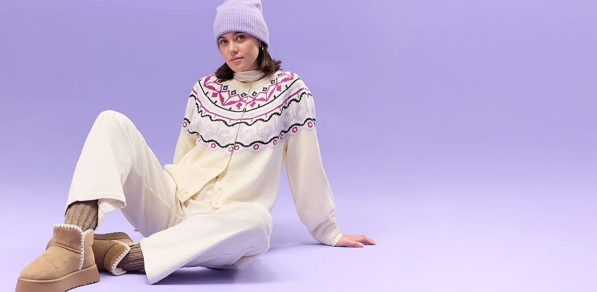 Maglione color crema con motivo nordico, pantaloni bianchi e berretto di maglia viola.