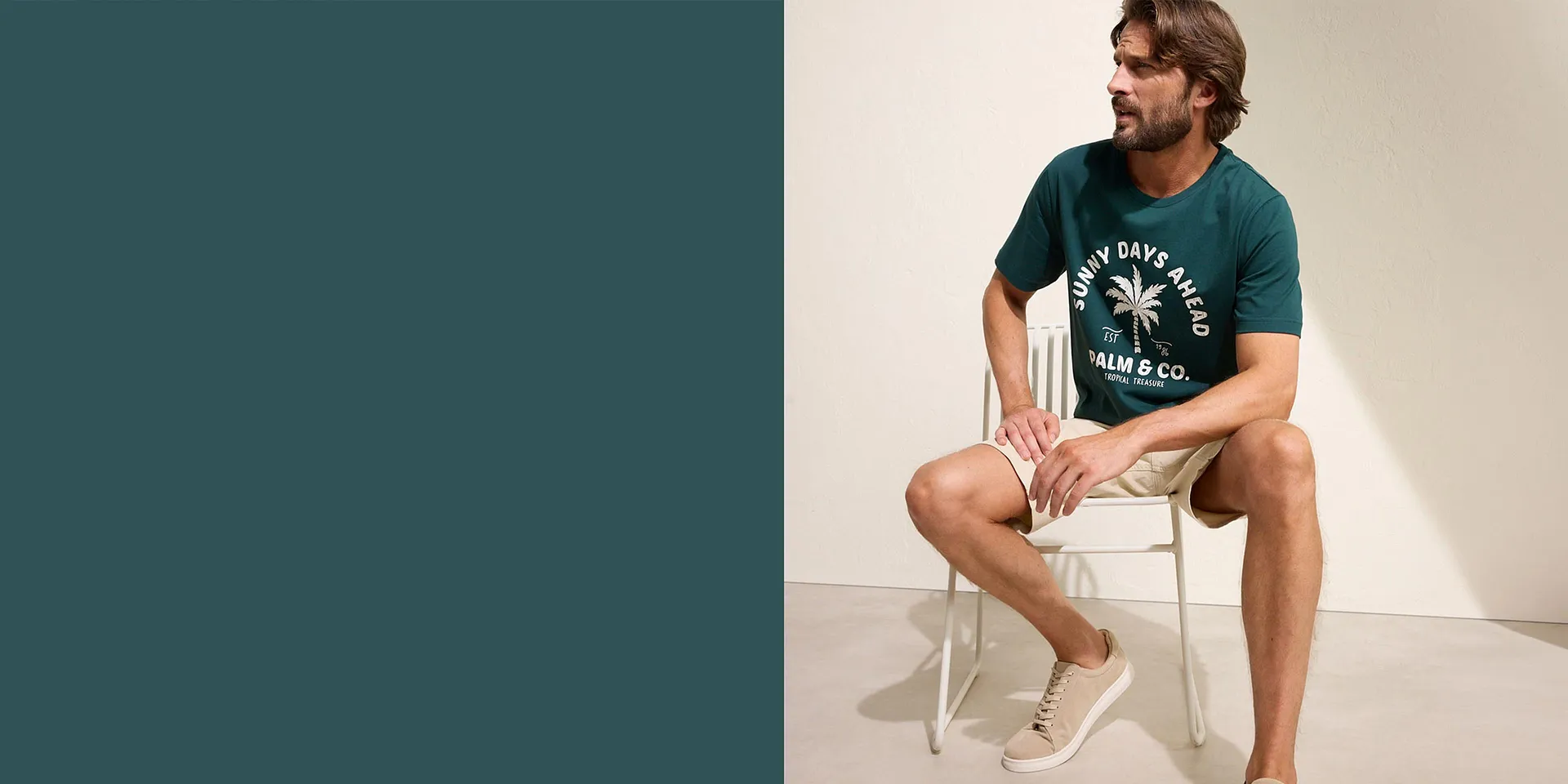 Un uomo indossa una maglietta verde con una stampa di palme, pantaloncini beige e sneaker chiare.