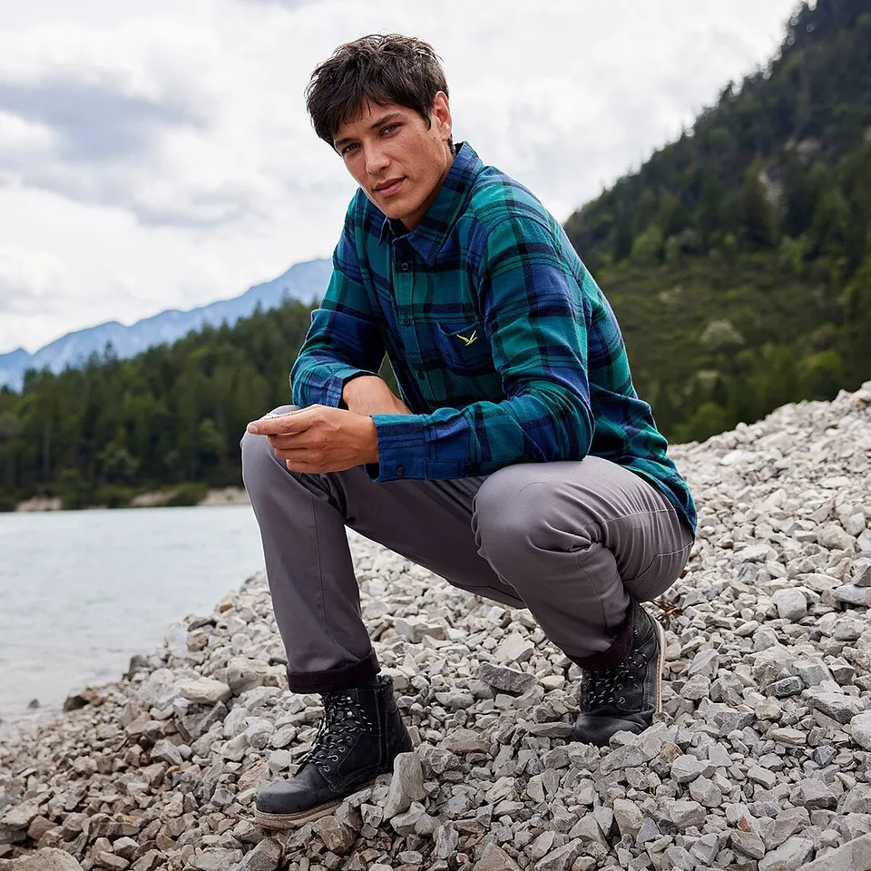 Un escursionista con camicia a quadri e pantaloni grigi è seduto sulla riva di un fiume in un paesaggio montuoso.