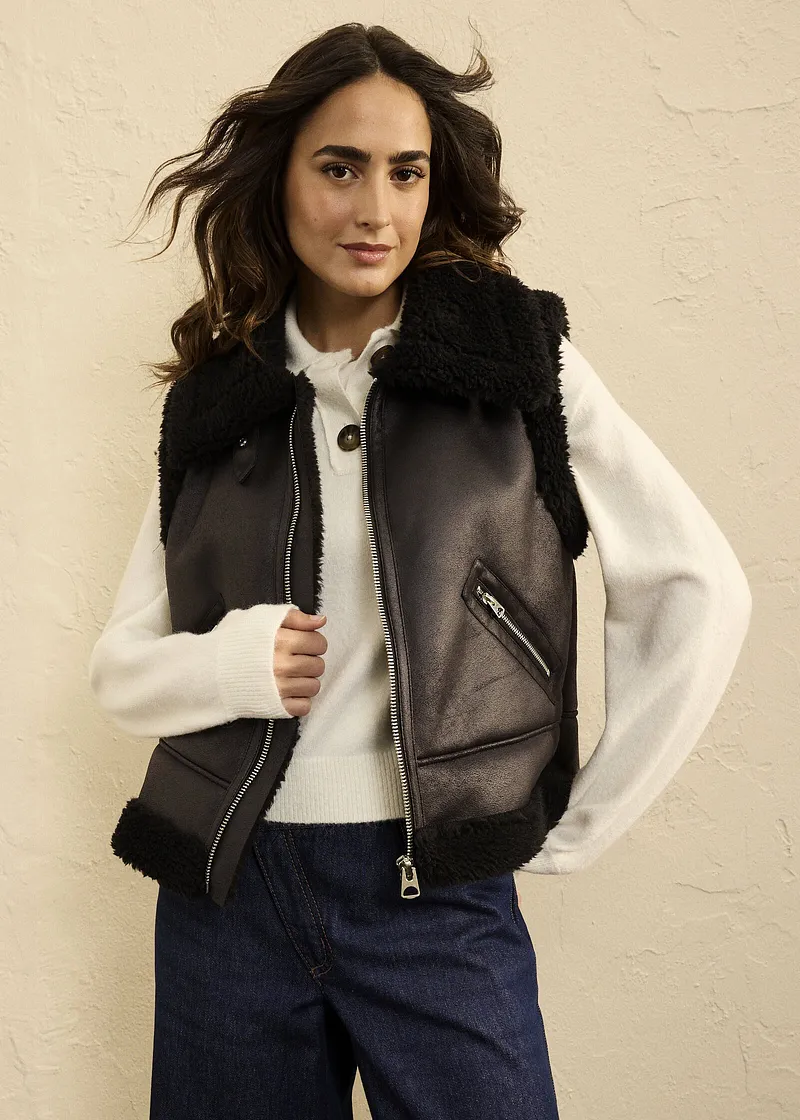Gilet in similpelle con collo di pelliccia sintetica, maglione bianco lavorato a maglia e jeans: look autunnale casual per le giornate fresche.