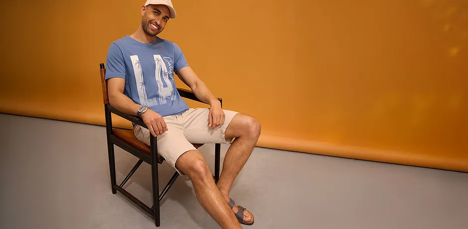 T-shirt blu con stampa, pantaloncini chiari, cappellino da baseball e sandali. 