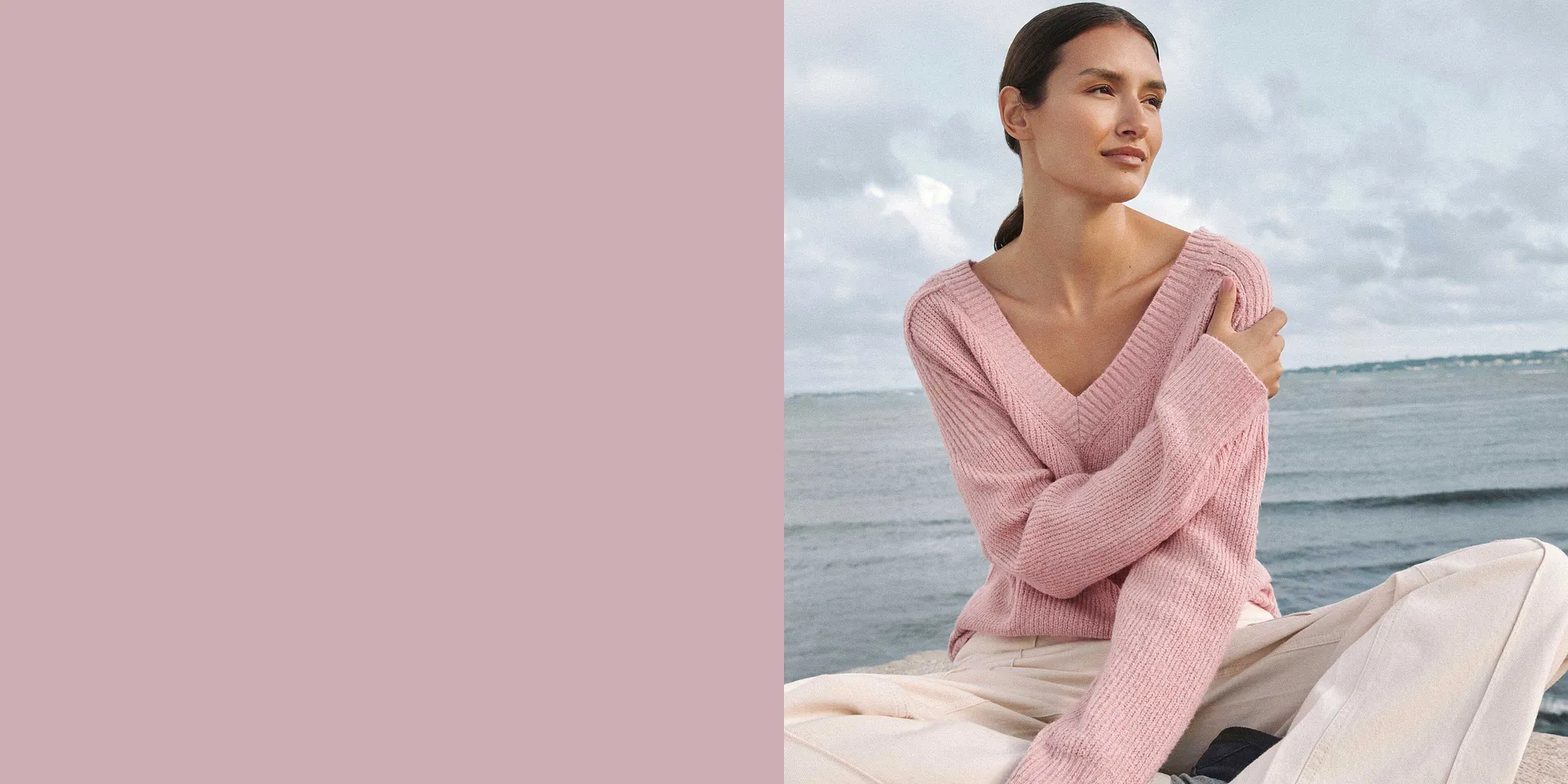 Un maglione rosa pallido con scollo a V impreziosisce la figura sullo sfondo del mare.