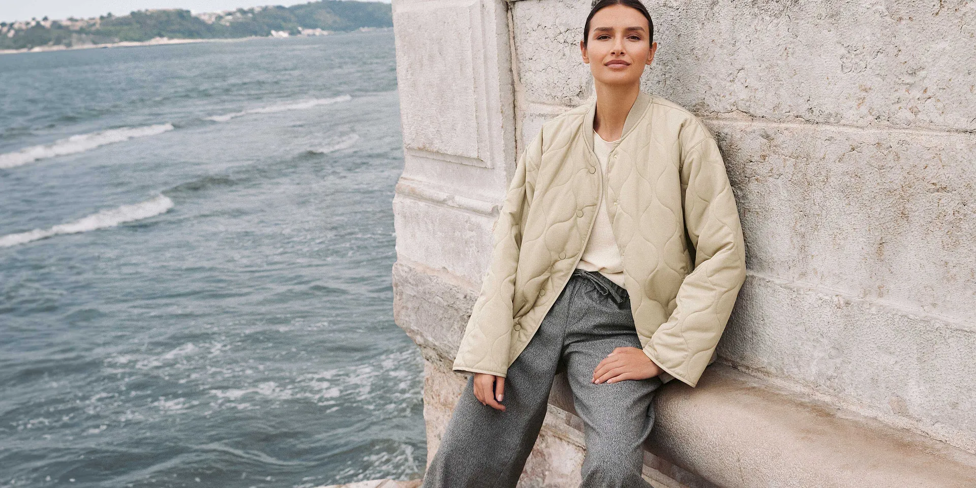 Look autunnale casual: giacca beige, jeans azzurri e maglia bianca in riva al mare.