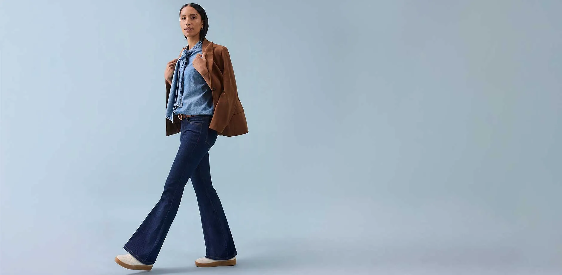 Una donna indossa jeans a zampa, un top senza maniche e una giacca marrone appoggiata sulle spalle.