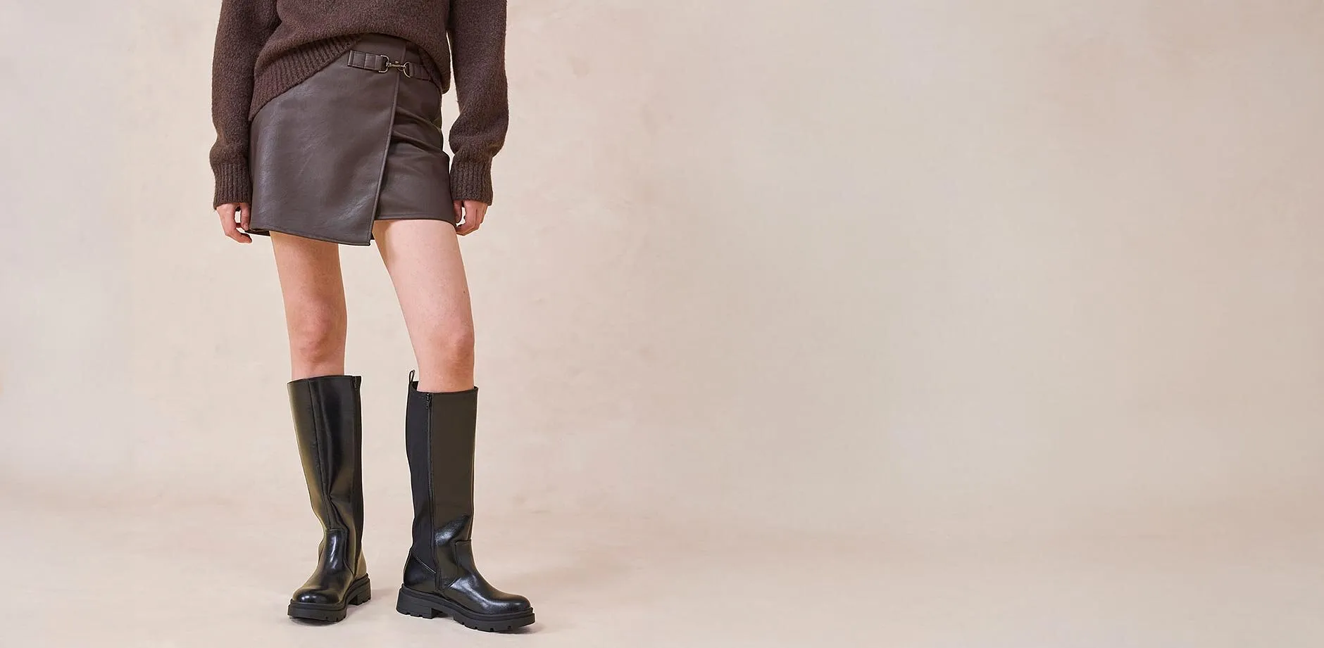 Gonna di pelle e stivali al ginocchio abbinati a un maglione marrone.