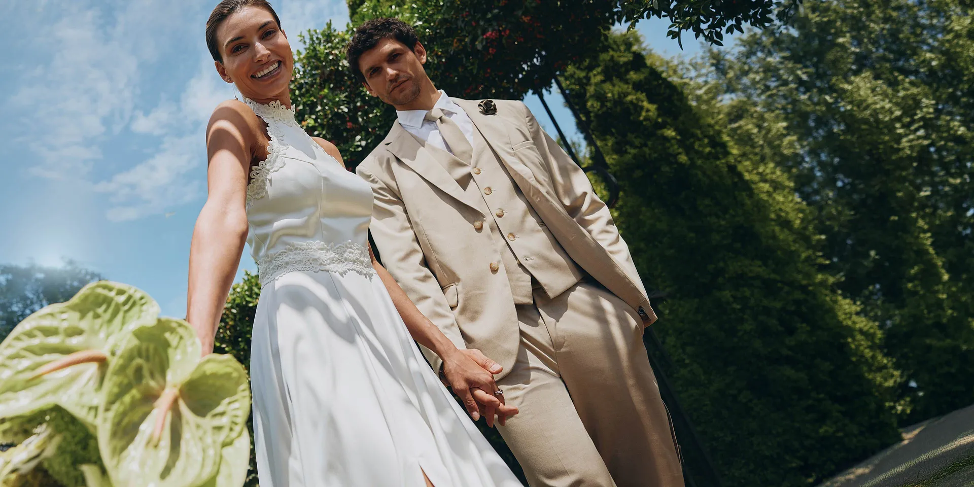 Una coppia di sposi elegante sotto il sole: lei in abito bianco, lui in abito beige con occhiali da sole.