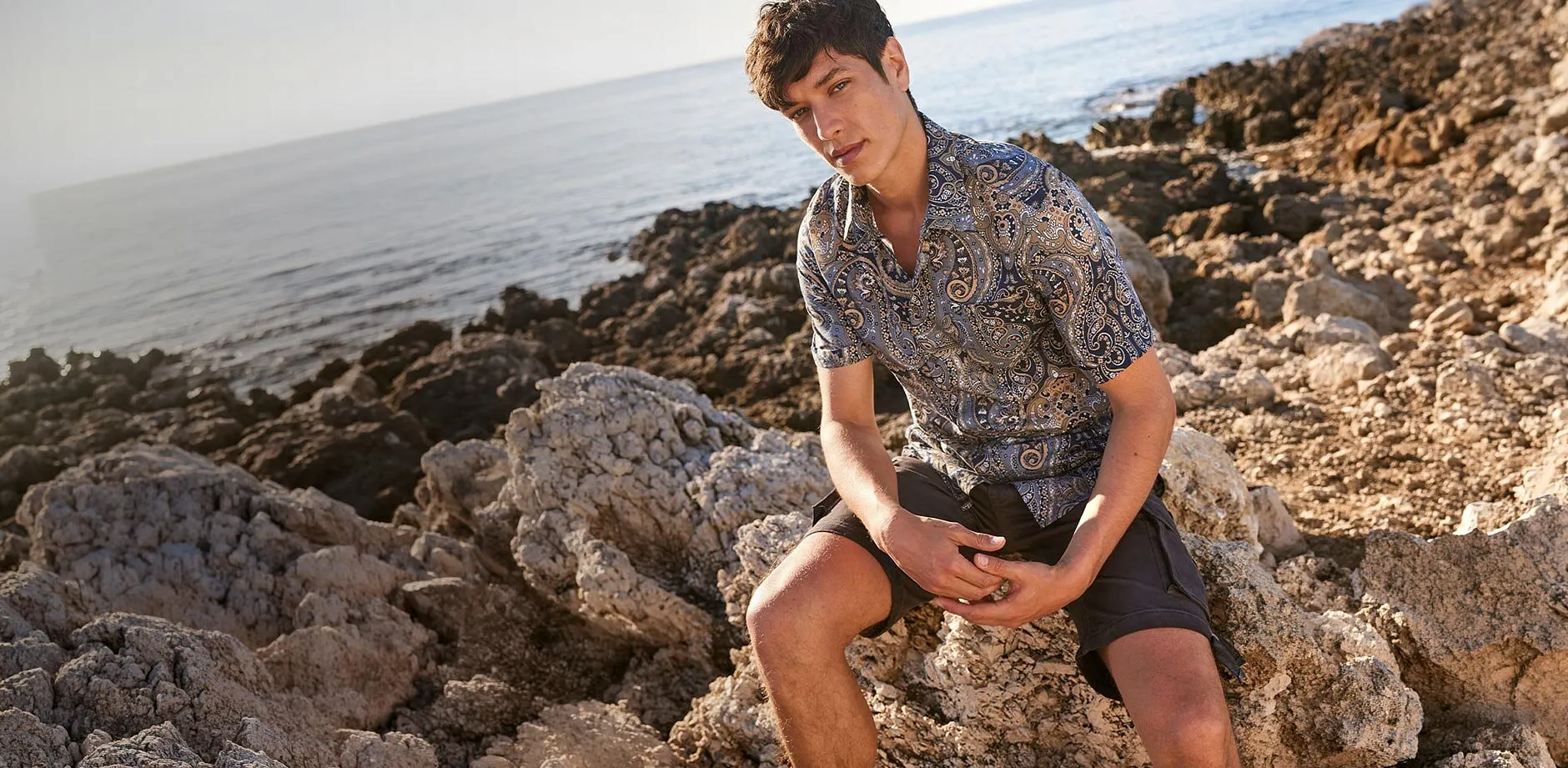 Un uomo indossa una camicia a fantasia e pantaloncini scuri su una spiaggia rocciosa.