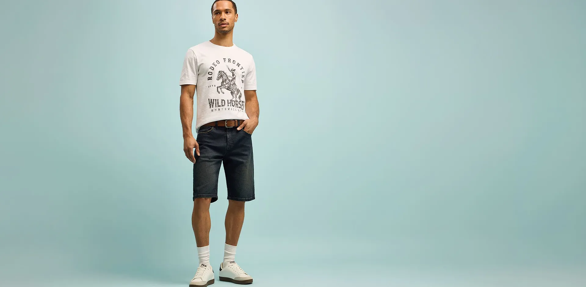 Un uomo indossa una maglietta bianca con una stampa e pantaloncini scuri, in una posa rilassata.