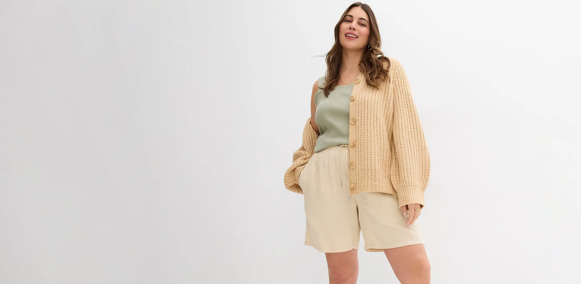 Una donna indossa un cardigan beige, un top verde chiaro e dei pantaloncini color crema.
