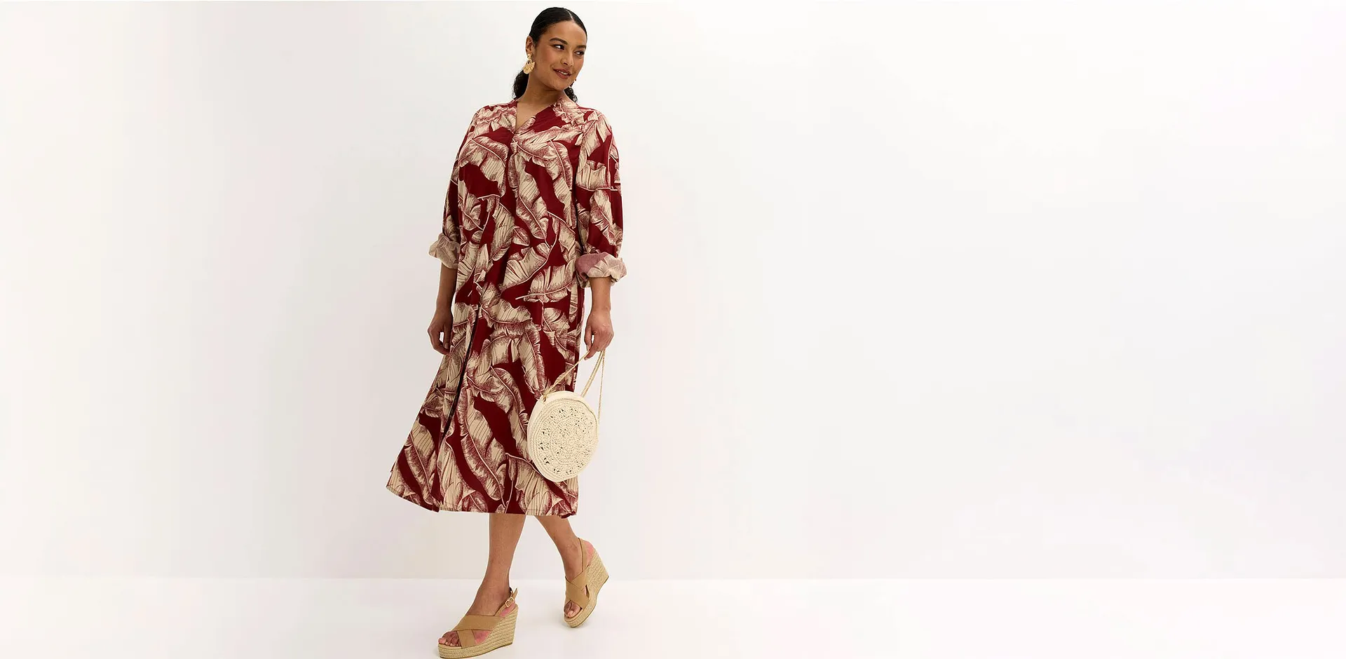 Abito midi con stampa botanica nei toni del rosso e del beige, abbinato a una borsa a mano intrecciata e a scarpe con tacco a zeppa.