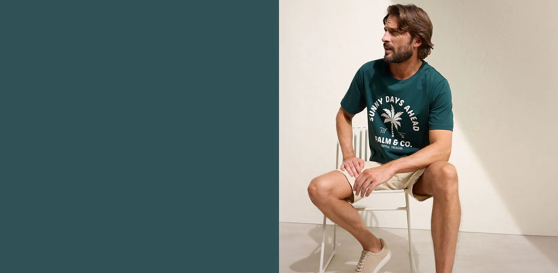Un uomo indossa una maglietta verde con una stampa di palme, pantaloncini beige e sneaker chiare.