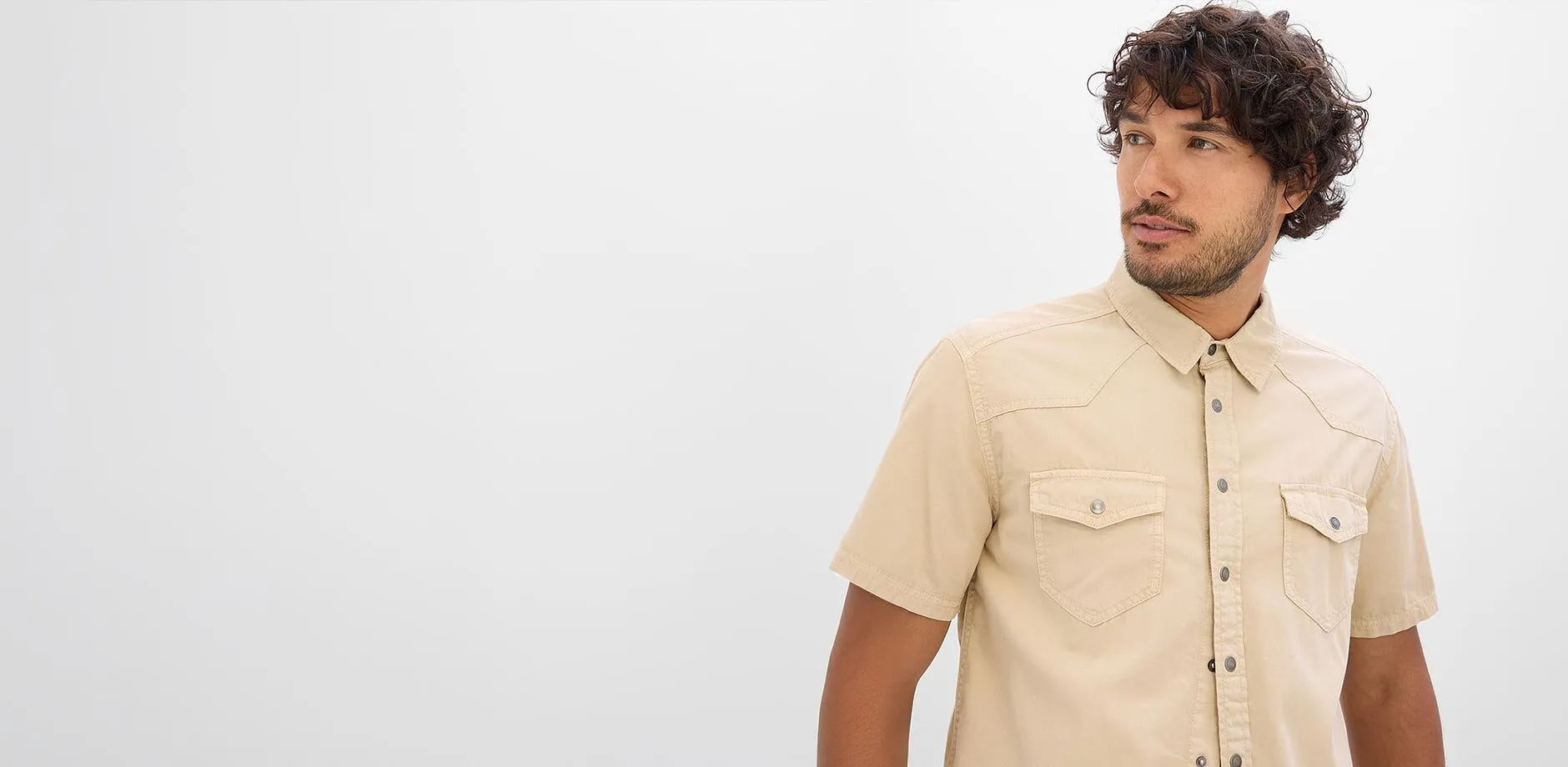 Un uomo indossa una camicia a maniche corte color beige chiaro con tasche sul petto e guarda di lato.
