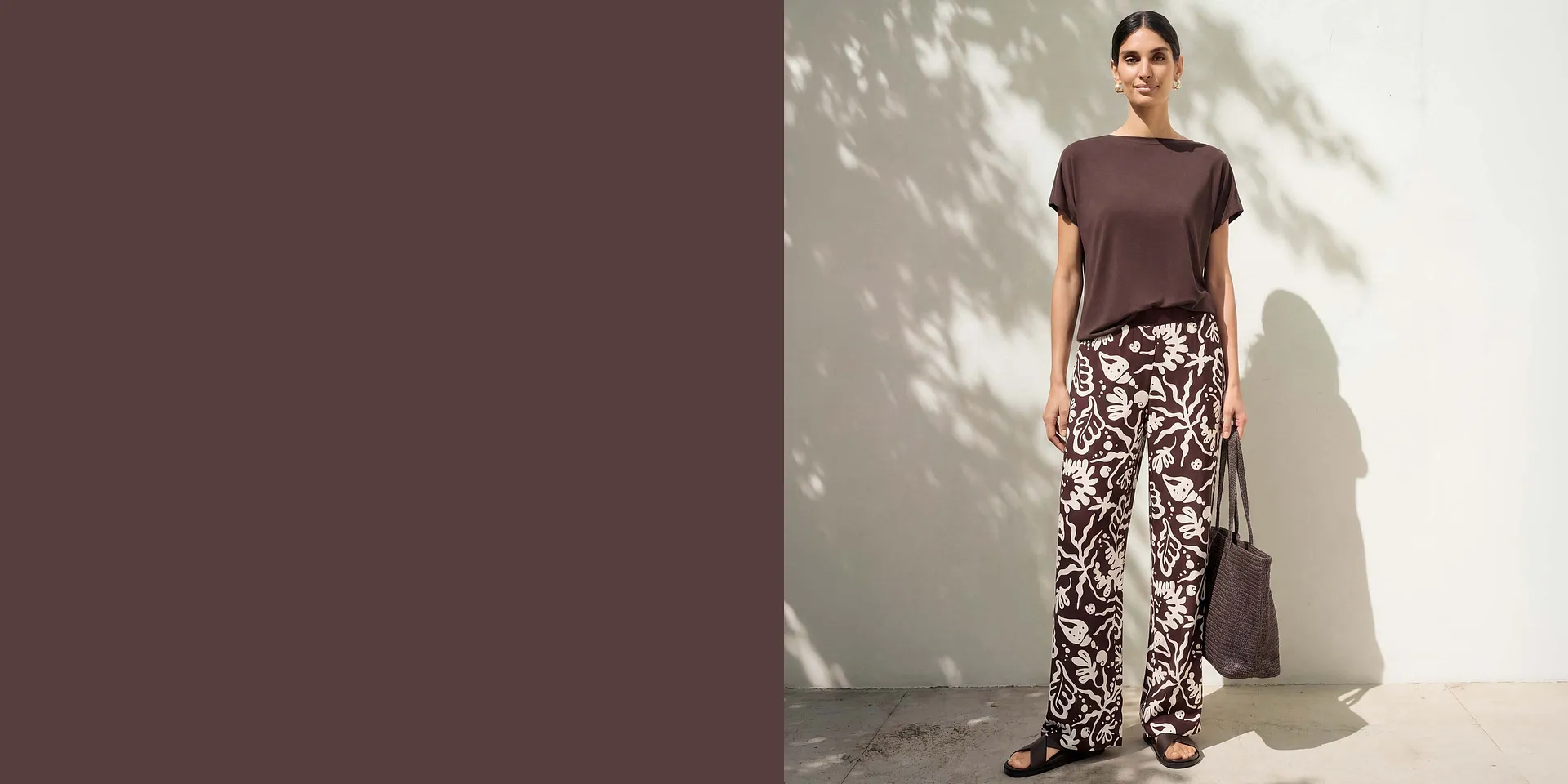 Maglia semplice marrone, pantaloni larghi a fantasia, abbinati a sandali scuri e una borsa.
