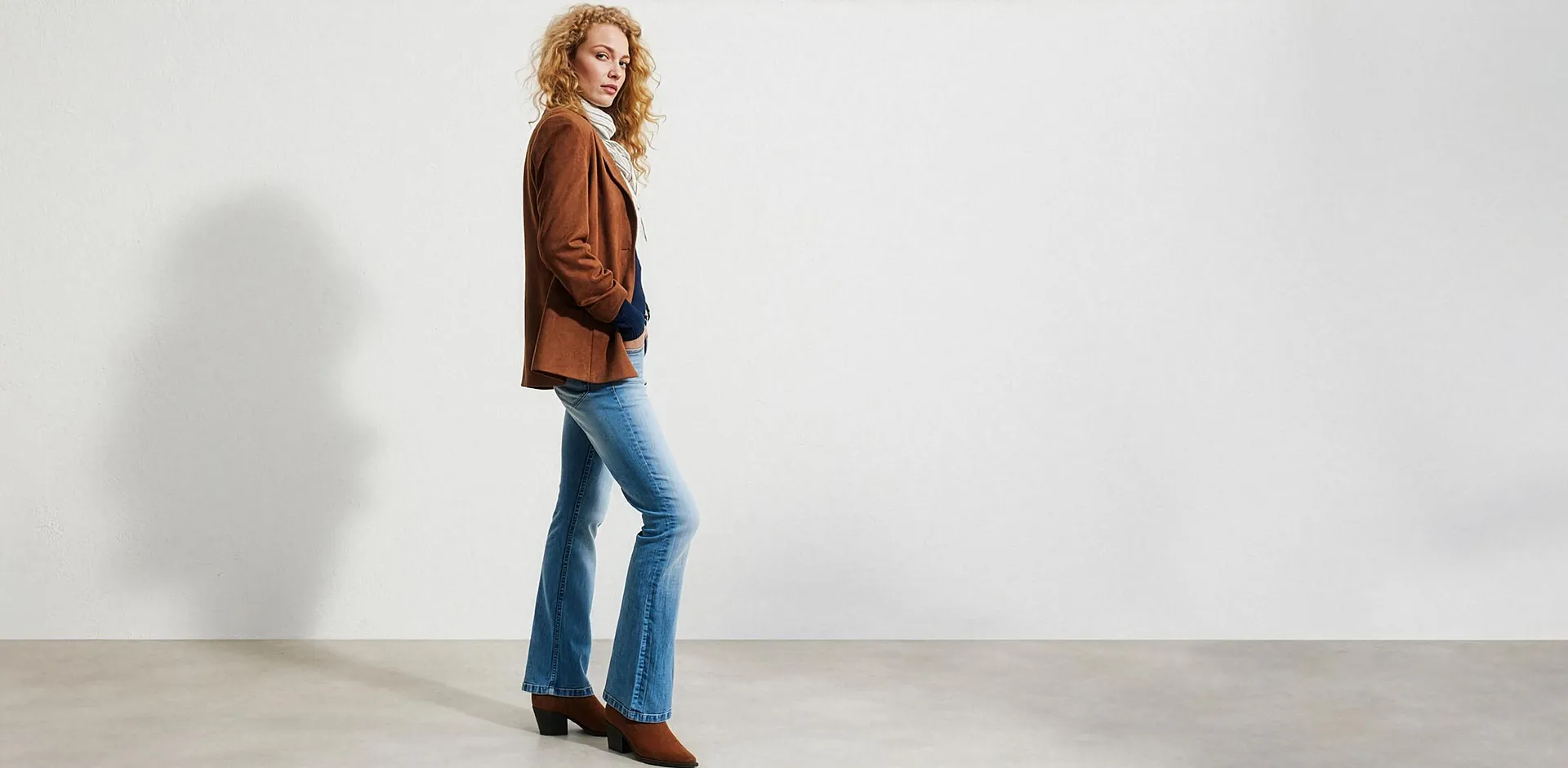 Giacca in pelle scamosciata marrone, jeans a zampa casual, maglione a collo alto bianco e stivaletti marroni.
