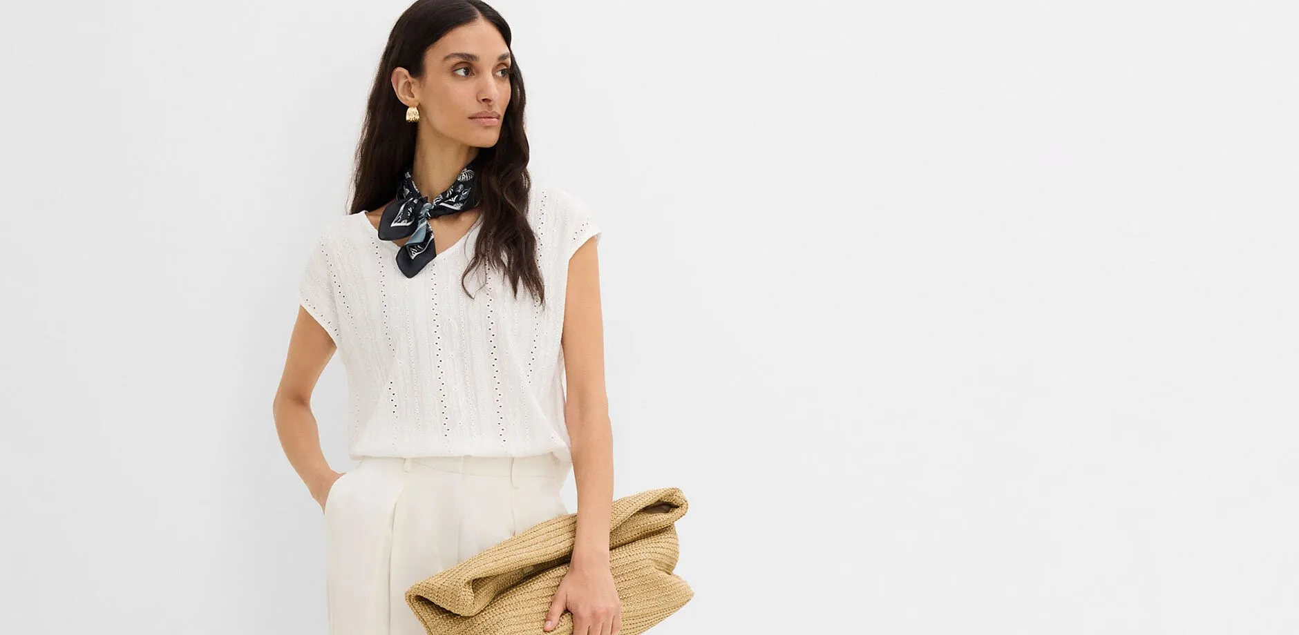 Maglia bianca con motivo traforato, pantaloni chiari, foulard scuro e borsa di paglia.