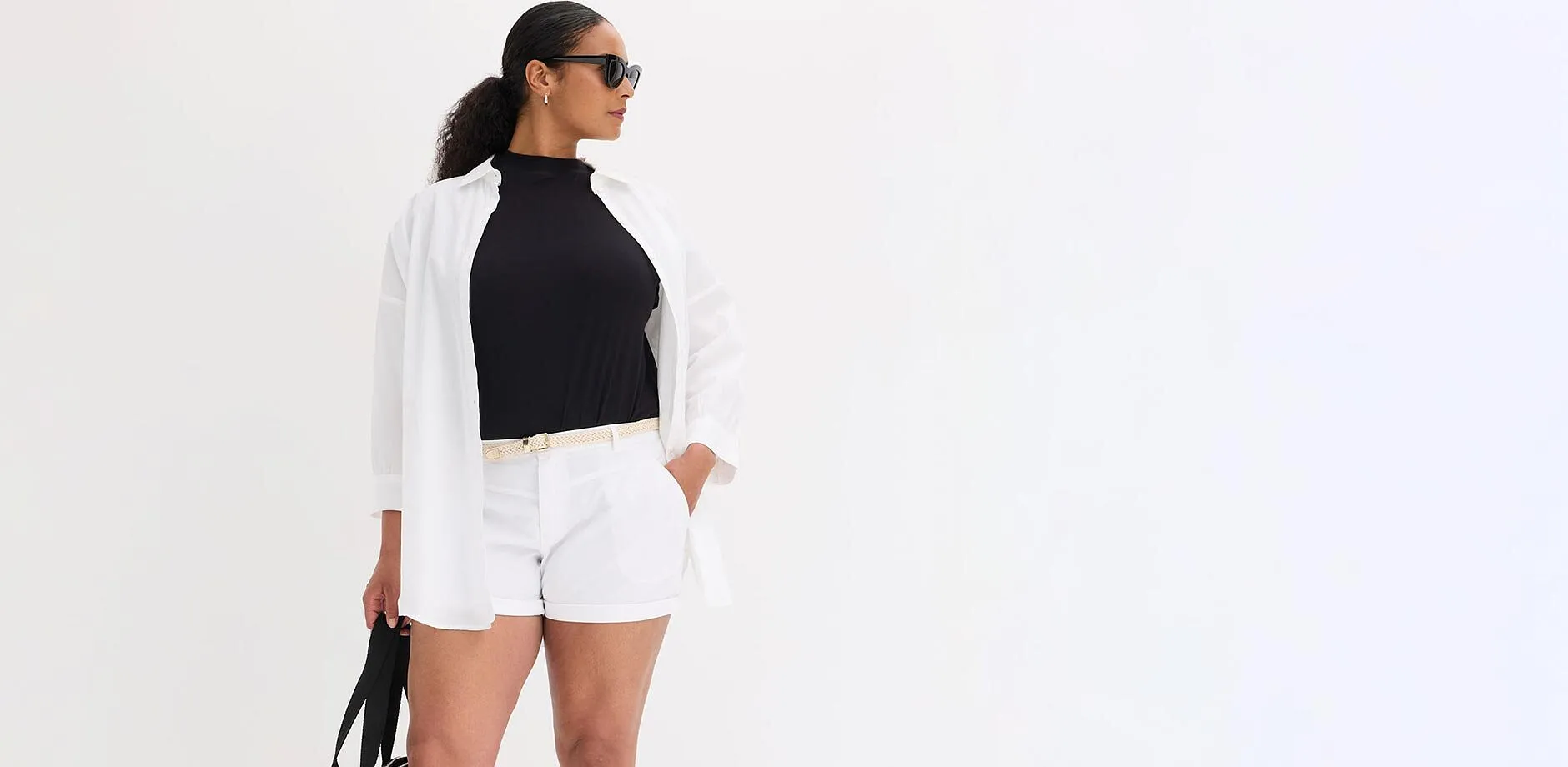Camicetta bianca aperta sopra un top nero, pantaloncini bianchi, cintura chiara, occhiali da sole.