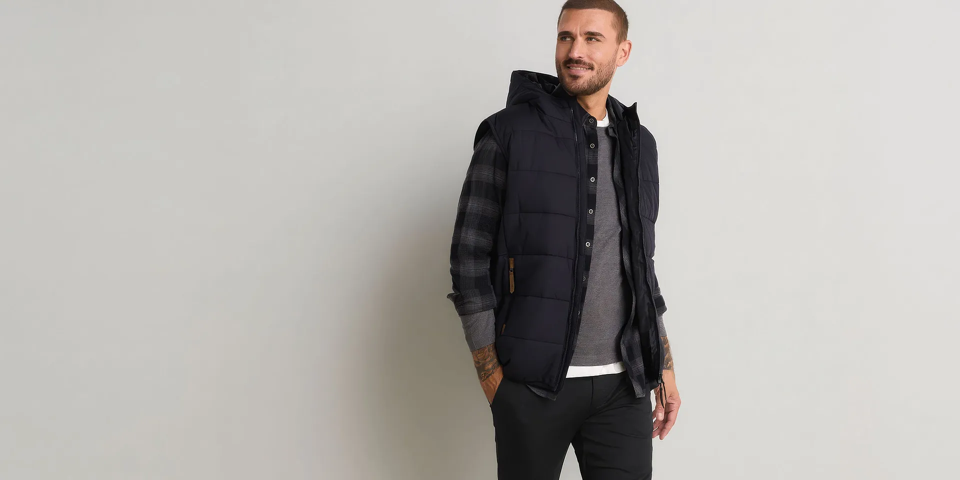 Gilet scuro trapuntato con cappuccio, camicia a quadri, maglietta grigia e pantaloni neri.