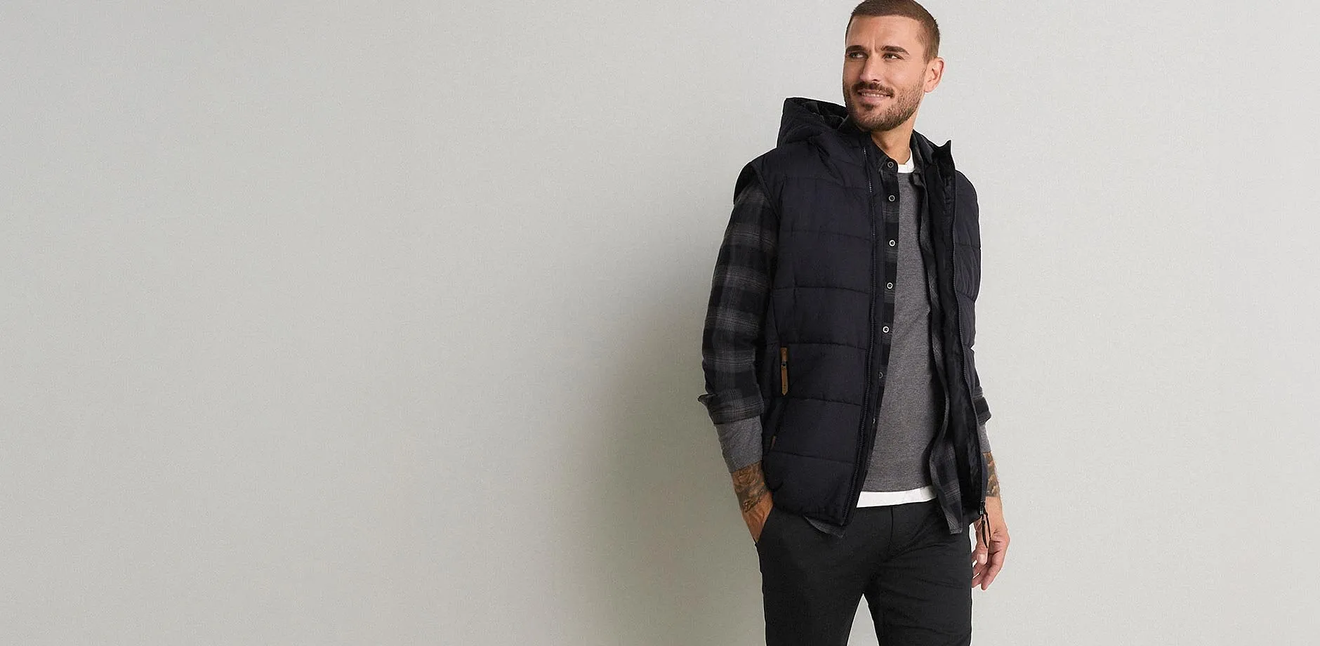 Gilet scuro trapuntato con cappuccio, camicia a quadri, maglietta grigia e pantaloni neri.