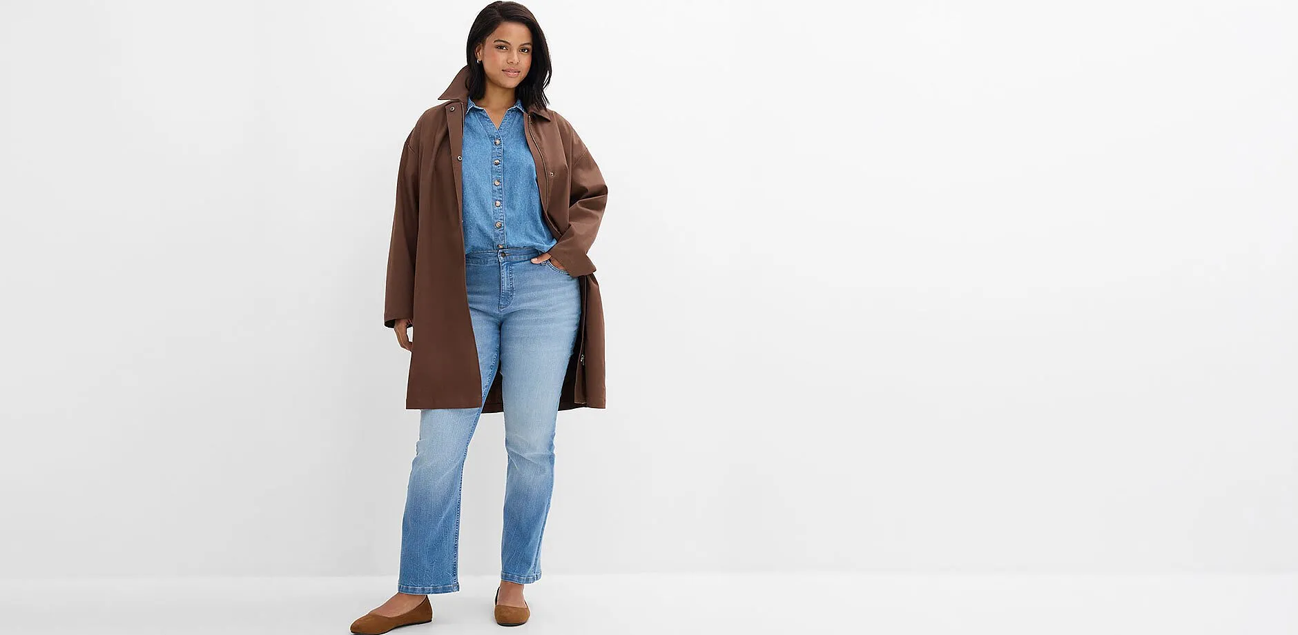 Un cappotto marrone sopra una camicia di jeans, jeans chiari e comode scarpe marroni.