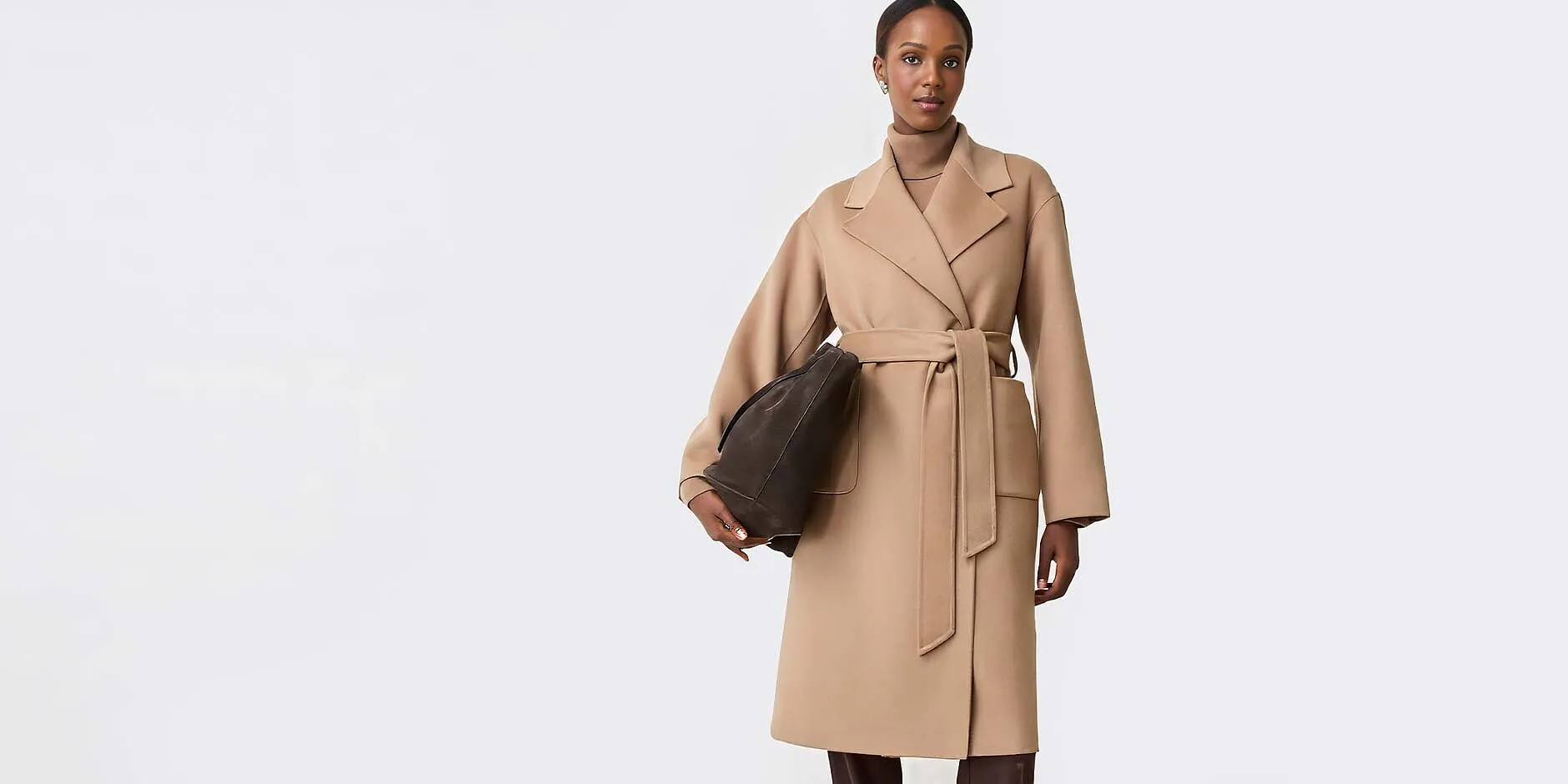 Elegante cappotto avvolgente color beige con cintura da annodare, maglione a collo alto e pantaloni scuri.