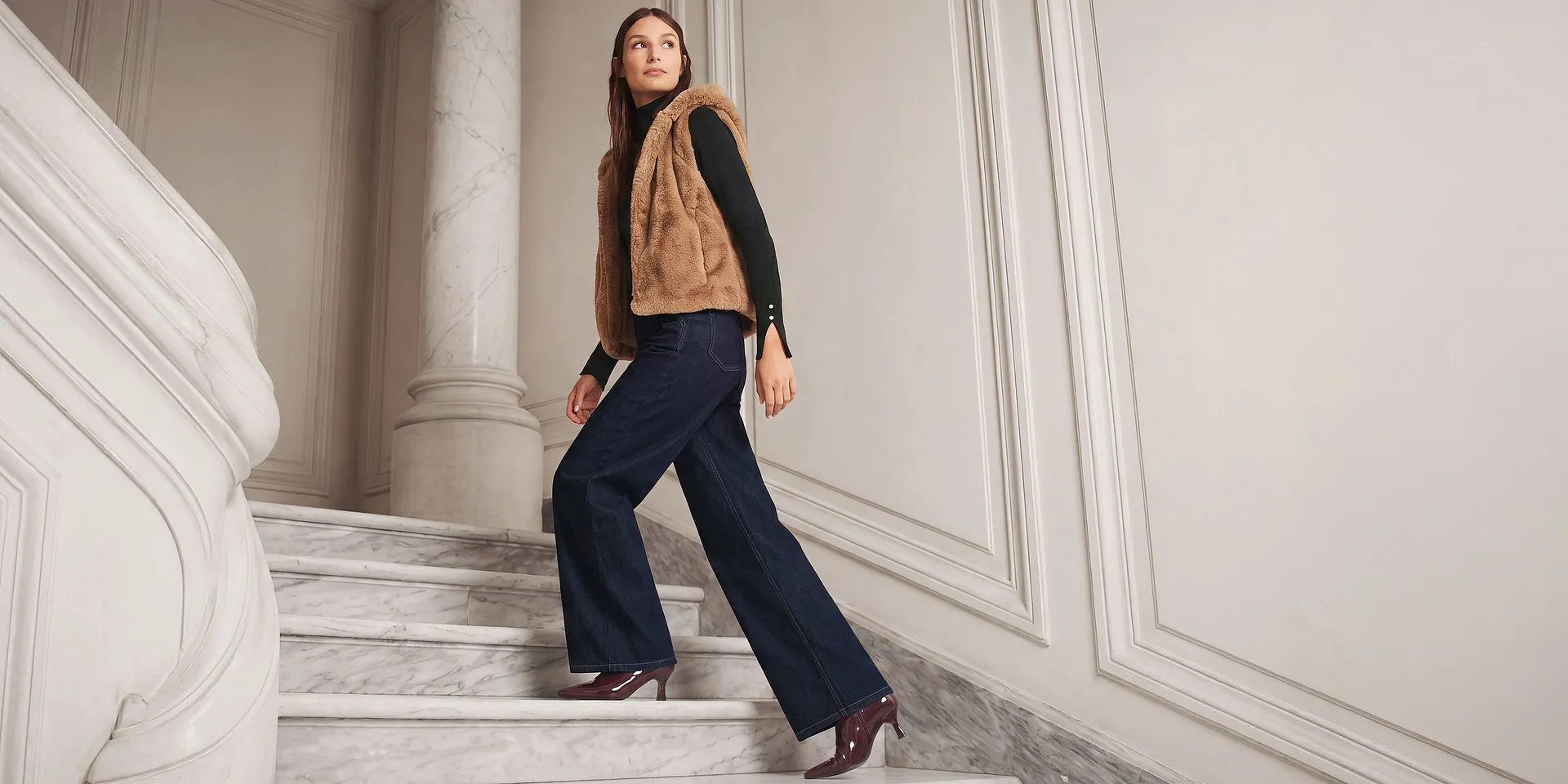 Gilet di eco-pelliccia marrone sopra un maglione a collo alto nero, abbinato a jeans scuri e stivaletti lucidi.