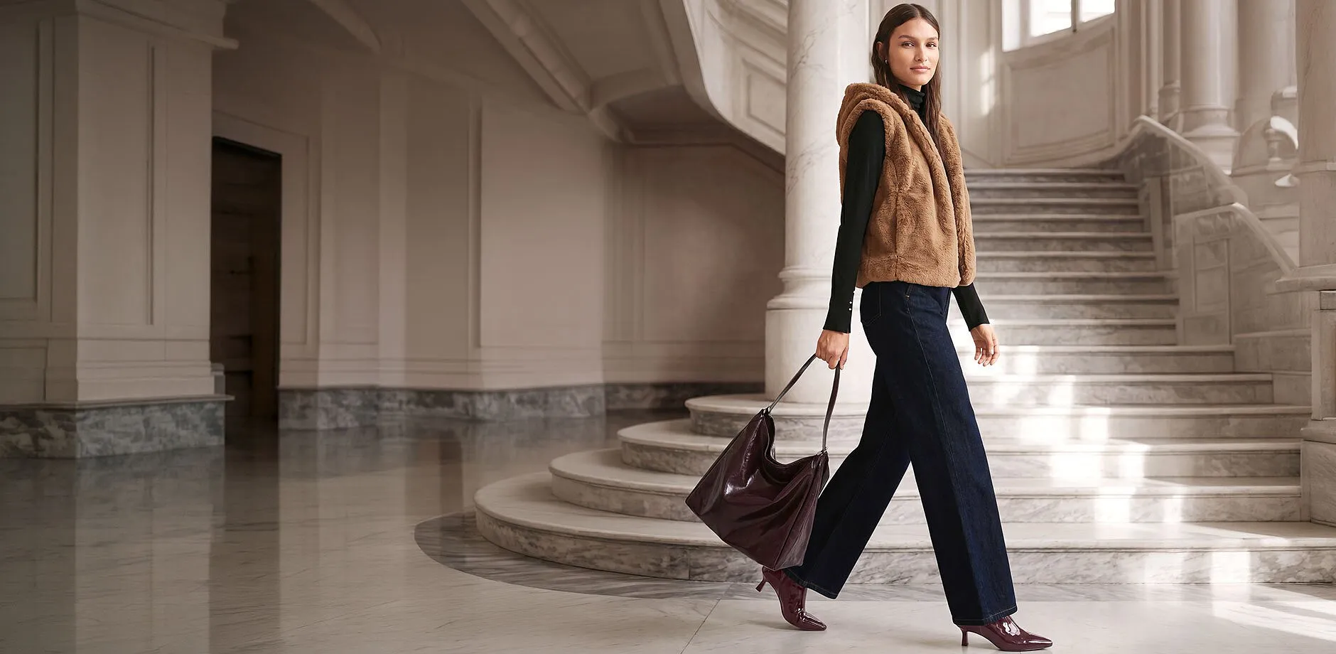 Gilet di eco-pelliccia marrone, maglione a collo alto nero, jeans larghi, borsa color bordeaux e stivaletti.