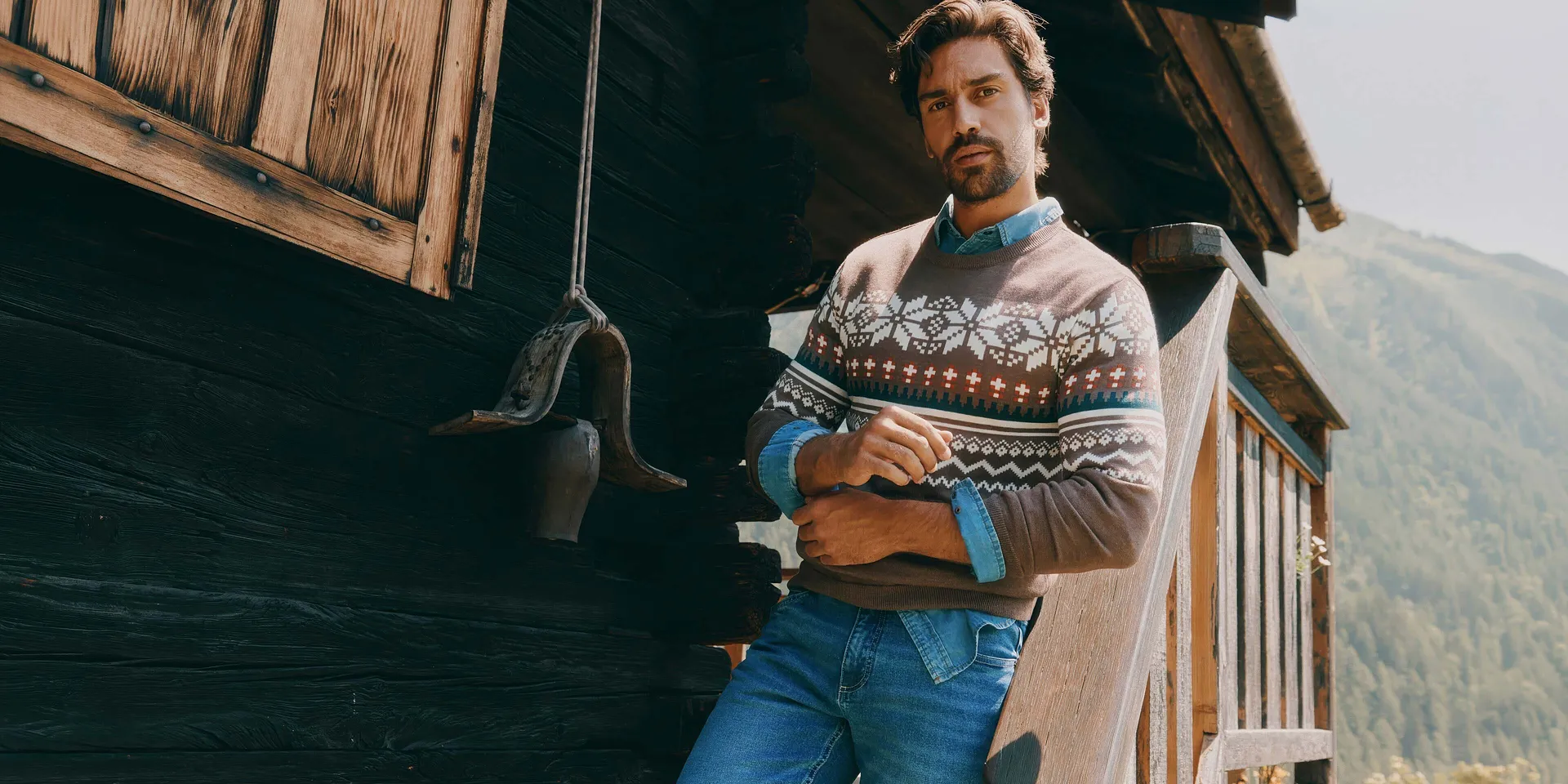 Indossa un maglione marrone a fantasia, una camicia di jeans e dei jeans blu.