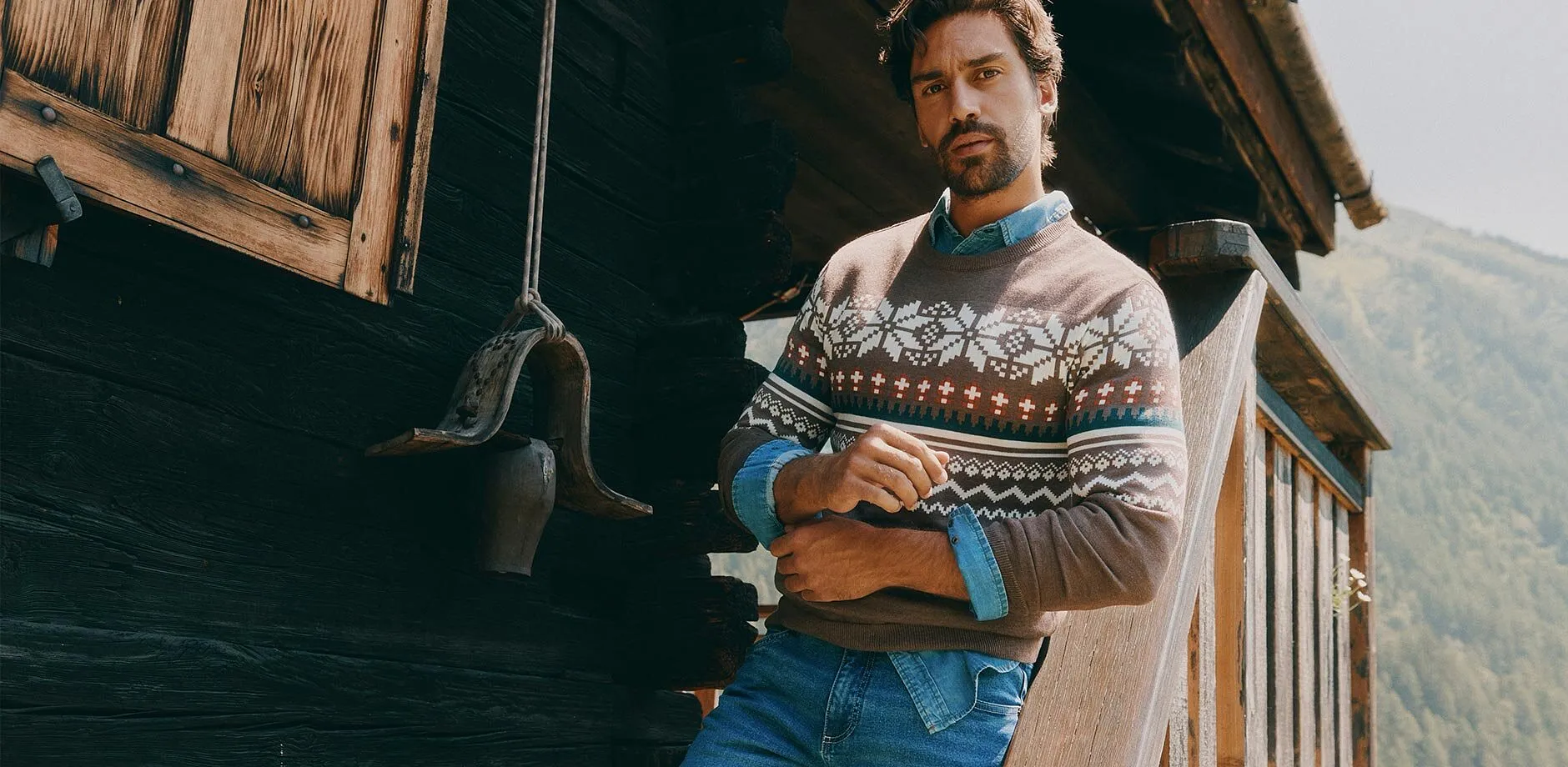 Indossa un maglione marrone a fantasia, una camicia di jeans e dei jeans blu.