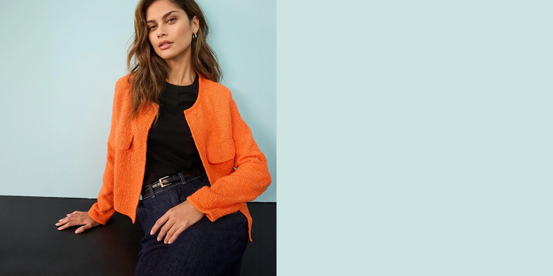 Cardigan arancione brillante sopra un top nero, abbinato a jeans e cintura elegante.