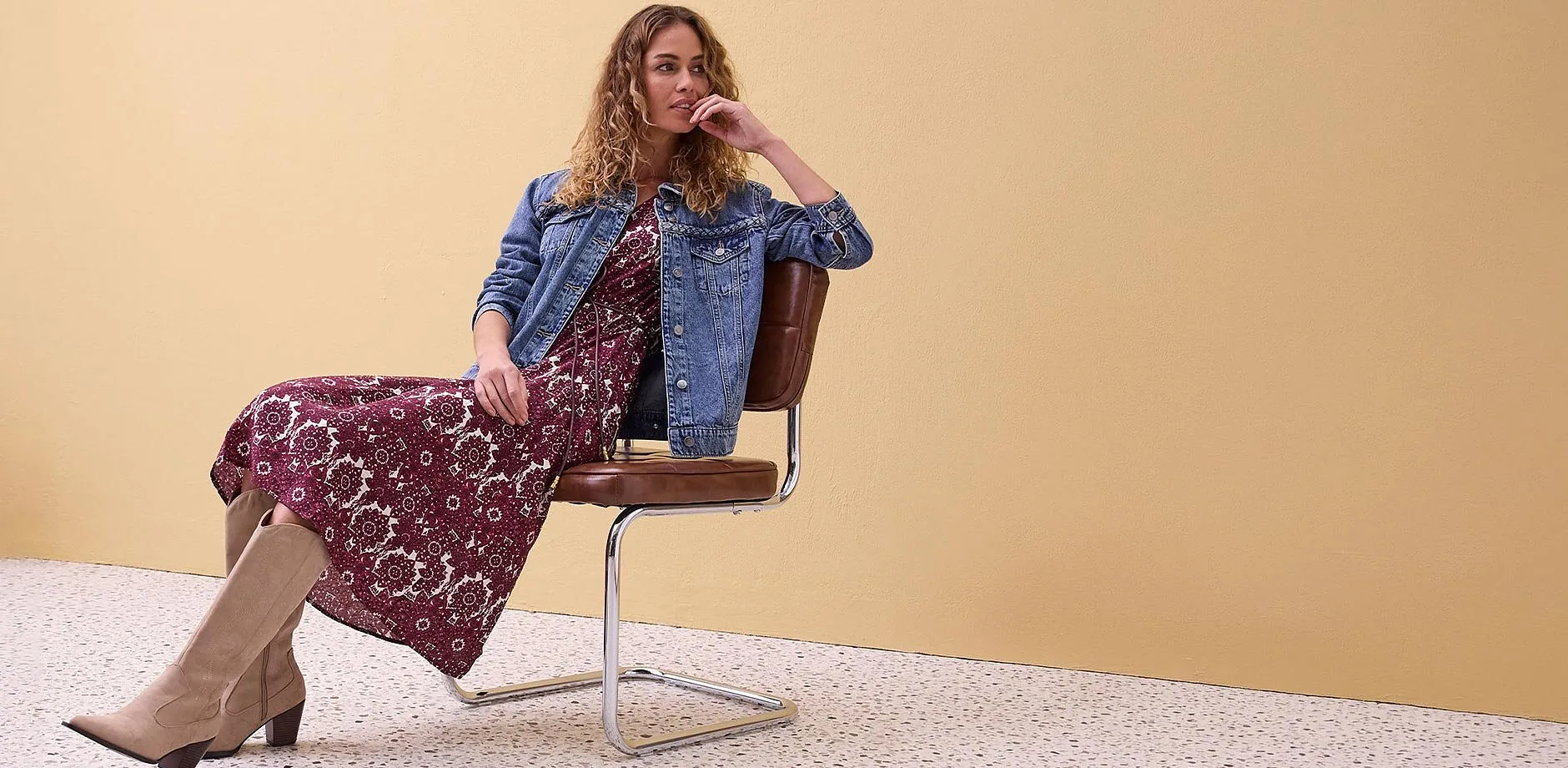 Giacca di jeans sopra un abito lungo a fiori, abbinata a stivali beige.