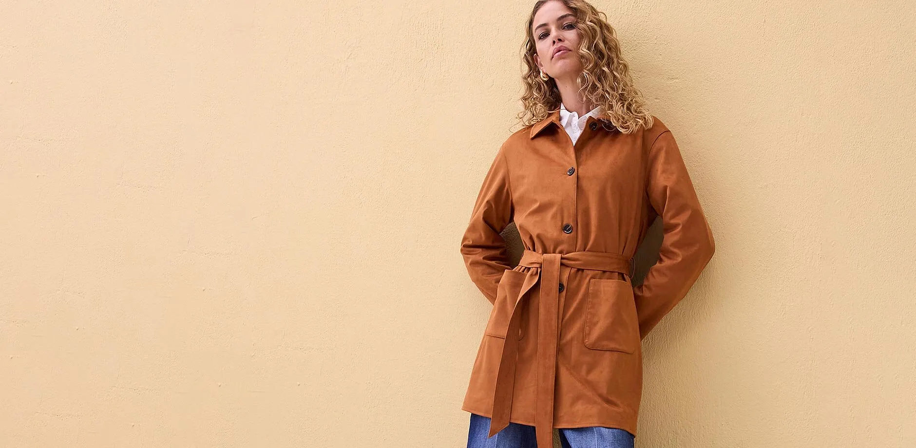 Cappotto color ruggine con cintura, abbinato a camicia bianca.