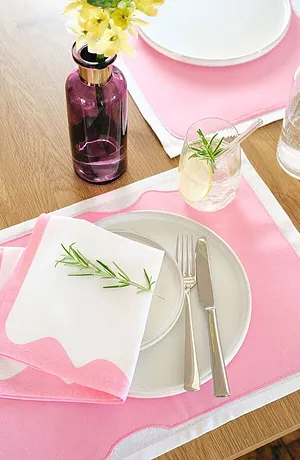 Una tovaglietta rosa tenue con motivo bianco decora il tavolo. Fiori freschi ed erbe aromatiche completano la tavola. 