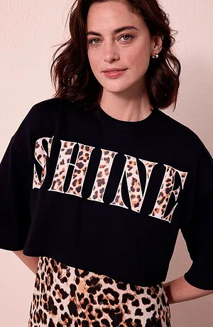 Donna con maglietta con scritta SHINE leopardata e gonna coordinata davanti a una parete chiara. 