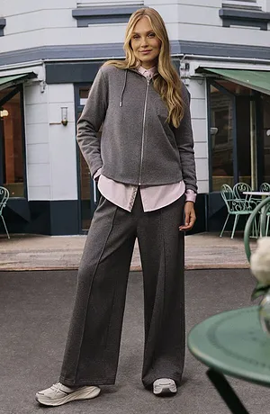 Felpa grigia con cappuccio sopra una camicetta rosa tenue, abbinata a pantaloni larghi grigi e sneakers bianche.