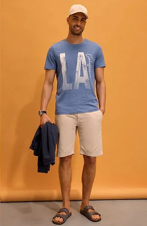 T-shirt blu con scritta "LA", pantaloncini chiari, cappello bianco e sandali. Look estivo casual.