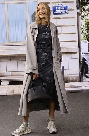Elegante trenchcoat sopra abito midi floreale, abbinato a sneakers bianche e borsetta nera.