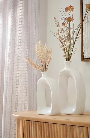 Tende chiare e leggere incorniciano eleganti vasi bianchi con fiori secchi su un armadio di legno.