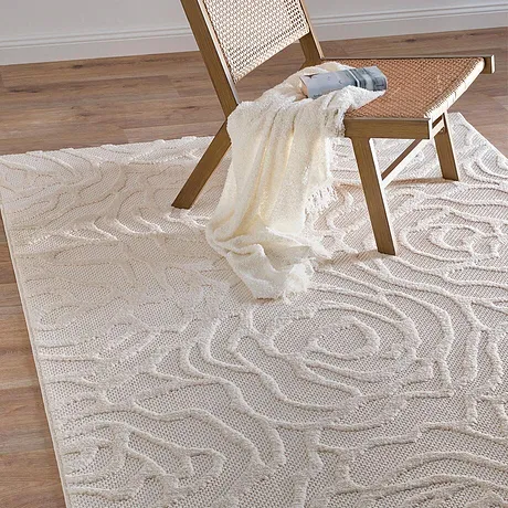 Una coperta color crema è posata con disinvoltura su una sedia in rattan su un elegante tappeto con motivi curvilinei. 