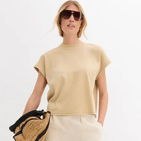 Outfit estivo con top beige a maniche corte, pantaloni bianchi, occhiali da sole e borsa di paglia. 