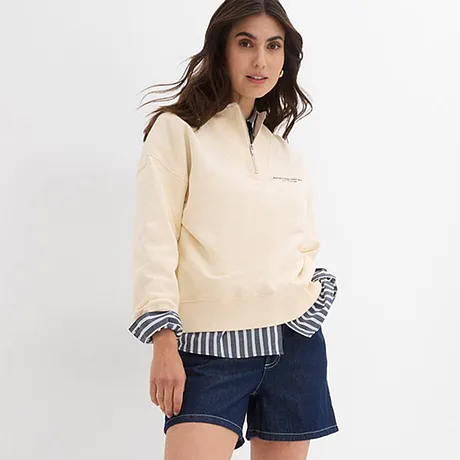 Pullover color crema sopra una camicia a righe, abbinato a shorts di jeans e sneakers bianche. 