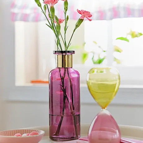 Vaso in vetro rosa con garofani rosa e clessidra decorativa rosa e gialla in un ambiente luminoso.