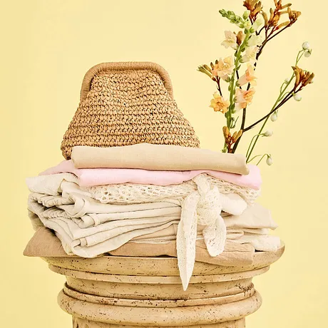 Accoglienti toni naturali: borsa di paglia, coperte delicate e un capo bianco su una colonna di legno.