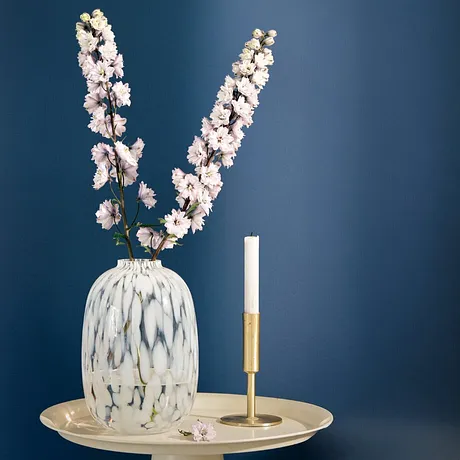 Elegante vaso bianco con delicati fiori di ciliegio su un vassoio dorato, accanto una candela sottile.