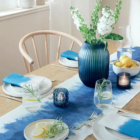 Un runner da tavolo blu decora il tavolo da pranzo, in contrasto con piatti bianchi e accessori colorati.