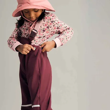 Look colorato da bambina: maglione rosa a fiori, pantaloni antipioggia bordeaux e cappello da sole color pastello.
