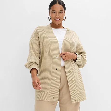 Cardigan oversize beige in maglia grossa, abbinato a top bianco e pantaloni beige chiaro. 