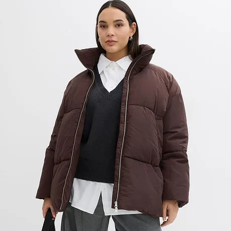 Giacca invernale marrone con collo alto, sotto maglione scuro e camicia bianca.