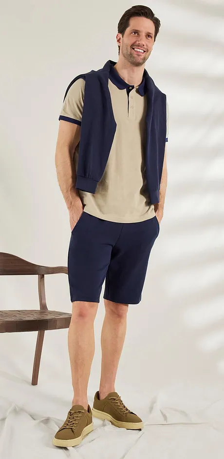 Look estivo casual: polo beige, pullover e pantaloncini al ginocchio blu scuro, sneakers marroni per uno stile rilassato. 