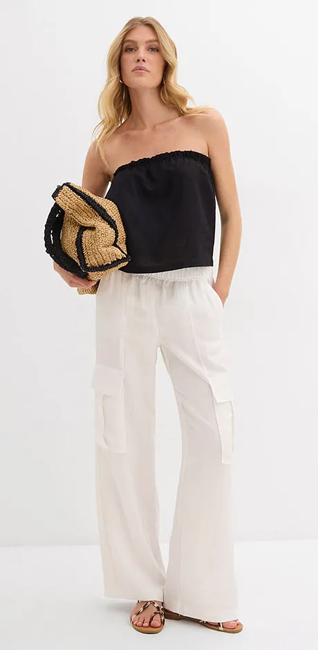 Outfit estivo con top nero senza spalle, pantaloni di lino bianchi e cappello intrecciato di paglia. 