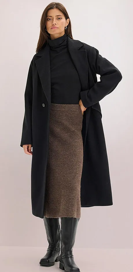 Elegante cappotto nero sopra un maglione a collo alto, abbinato a una gonna midi marrone e stivali di pelle al ginocchio.